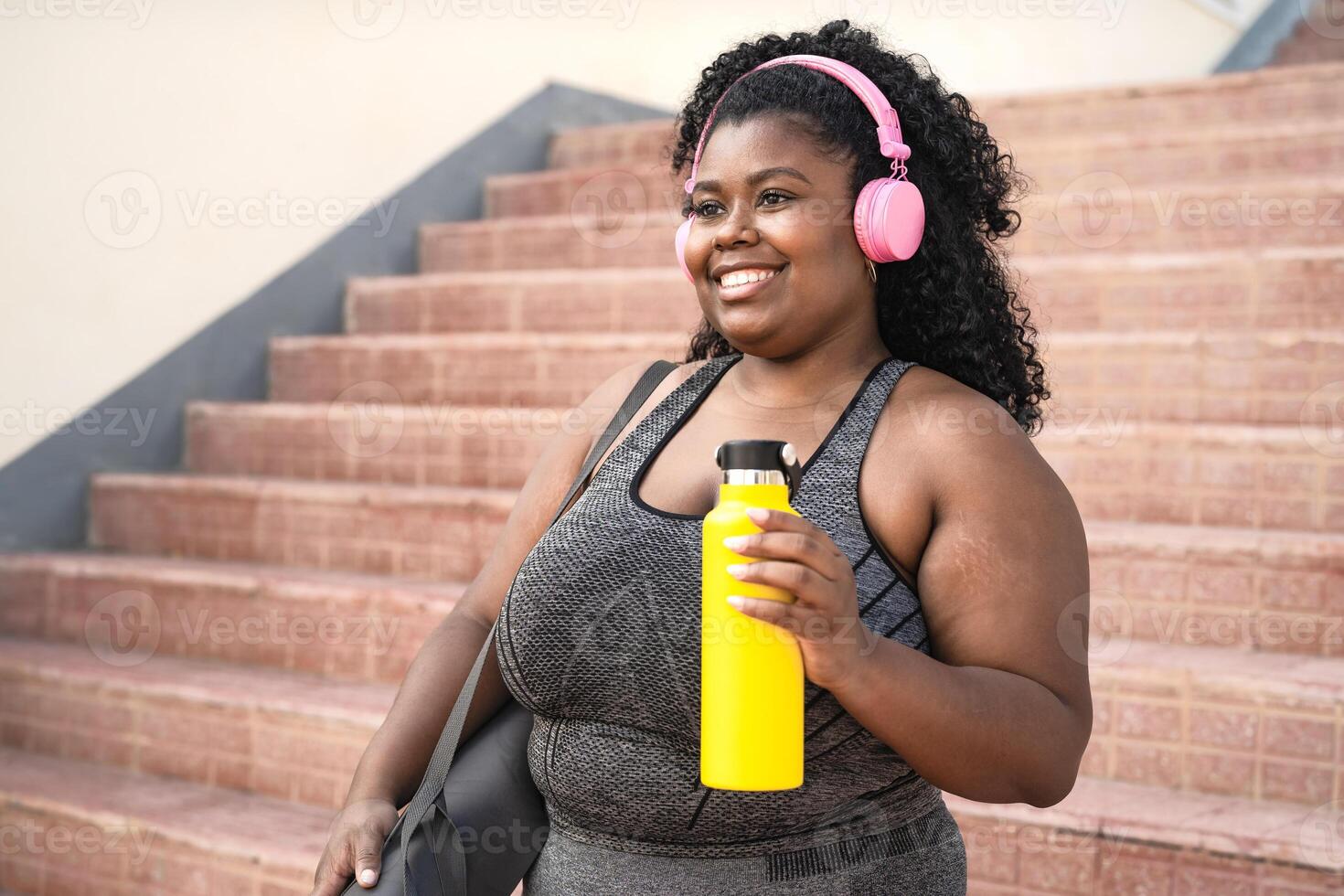 Happy plus size African woman doing workout routine while listening music with wireless headphones outdoor photo
