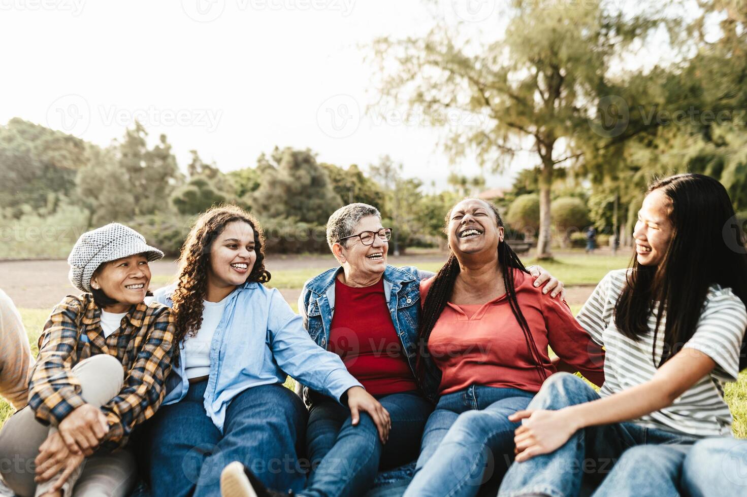 Happy multi generational group of women with different ethnicities having fun sitting on grass in a public park - Females empowerment concept photo