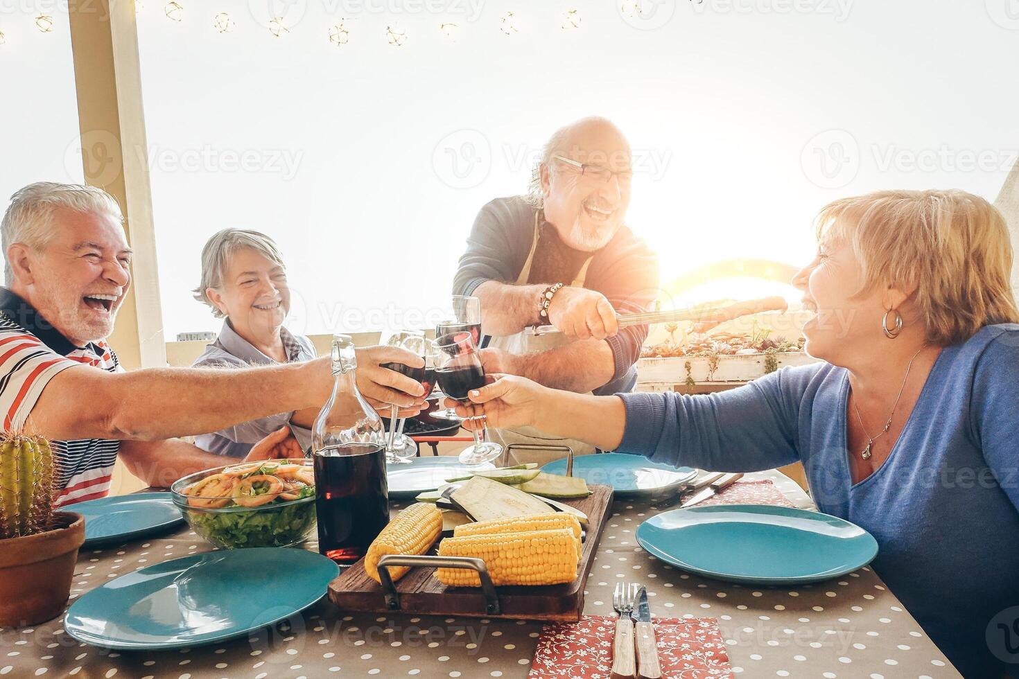 Happy senior friends having fun cheering with red wine at barbecue in terrace outdoor - Older people dining with grill meat and laughing together - Friendship and elderly lifestyle concept photo