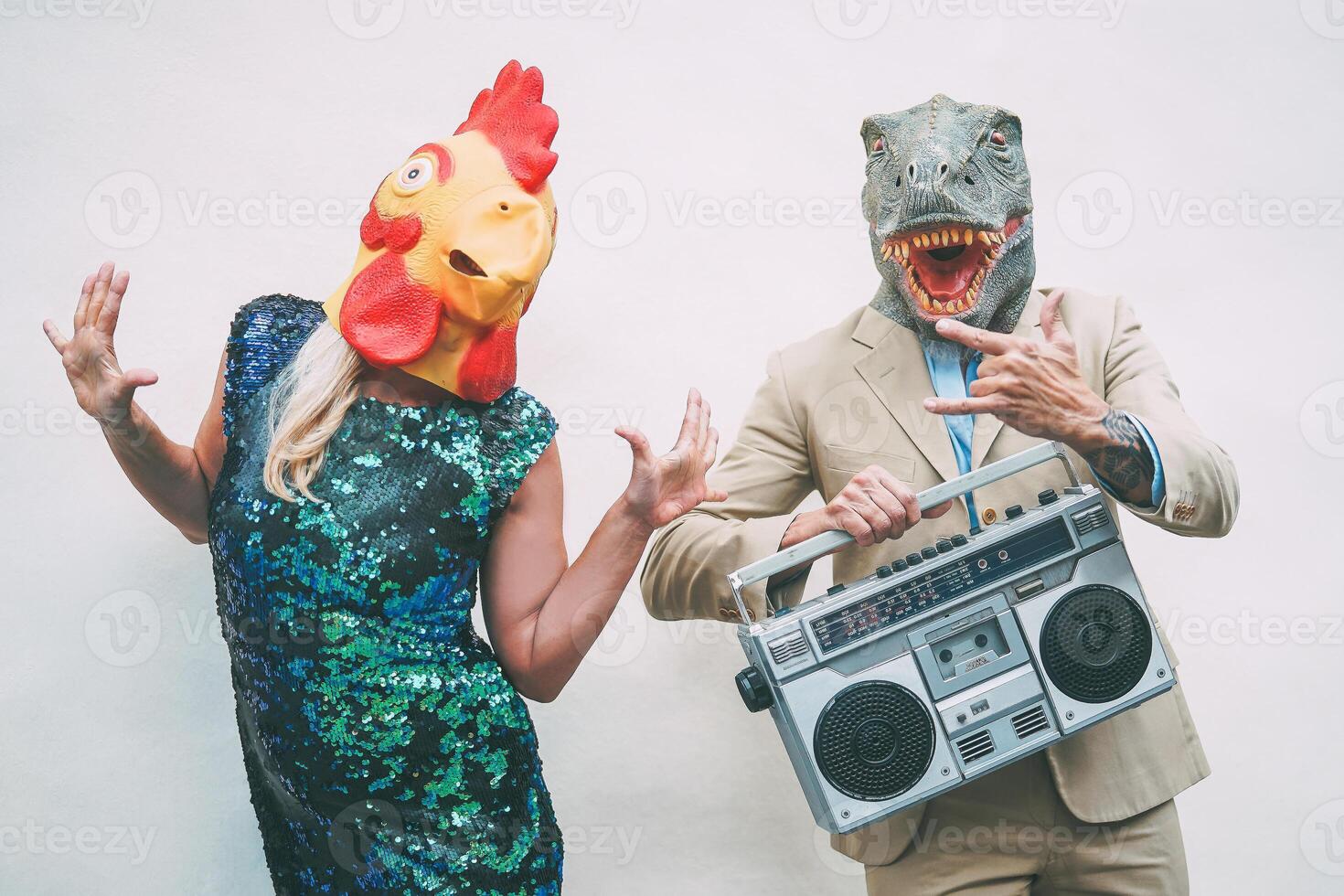 Crazy senior couple wearing chicken and t-rex mask while dancing outdoor - Mature trendy people having fun celebrating and listening music with boombox - Absurd concept of masquerade funny holidays photo