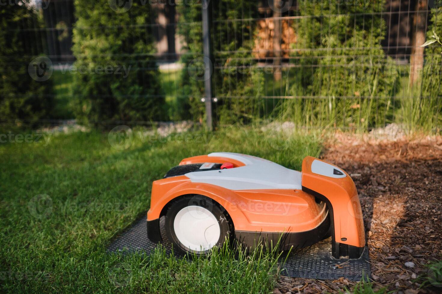 An orange robotic lawn mower stands on the base and is charged from electricity in the courtyard photo