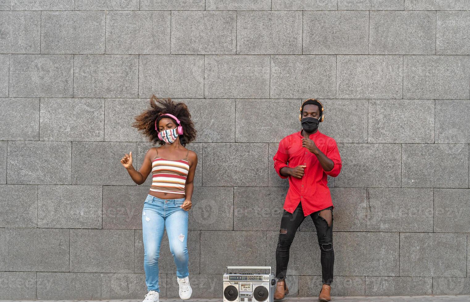 joven africano personas vistiendo cara máscara y bailando al aire libre mientras escuchando a música con inalámbrico auriculares y Clásico boombox foto