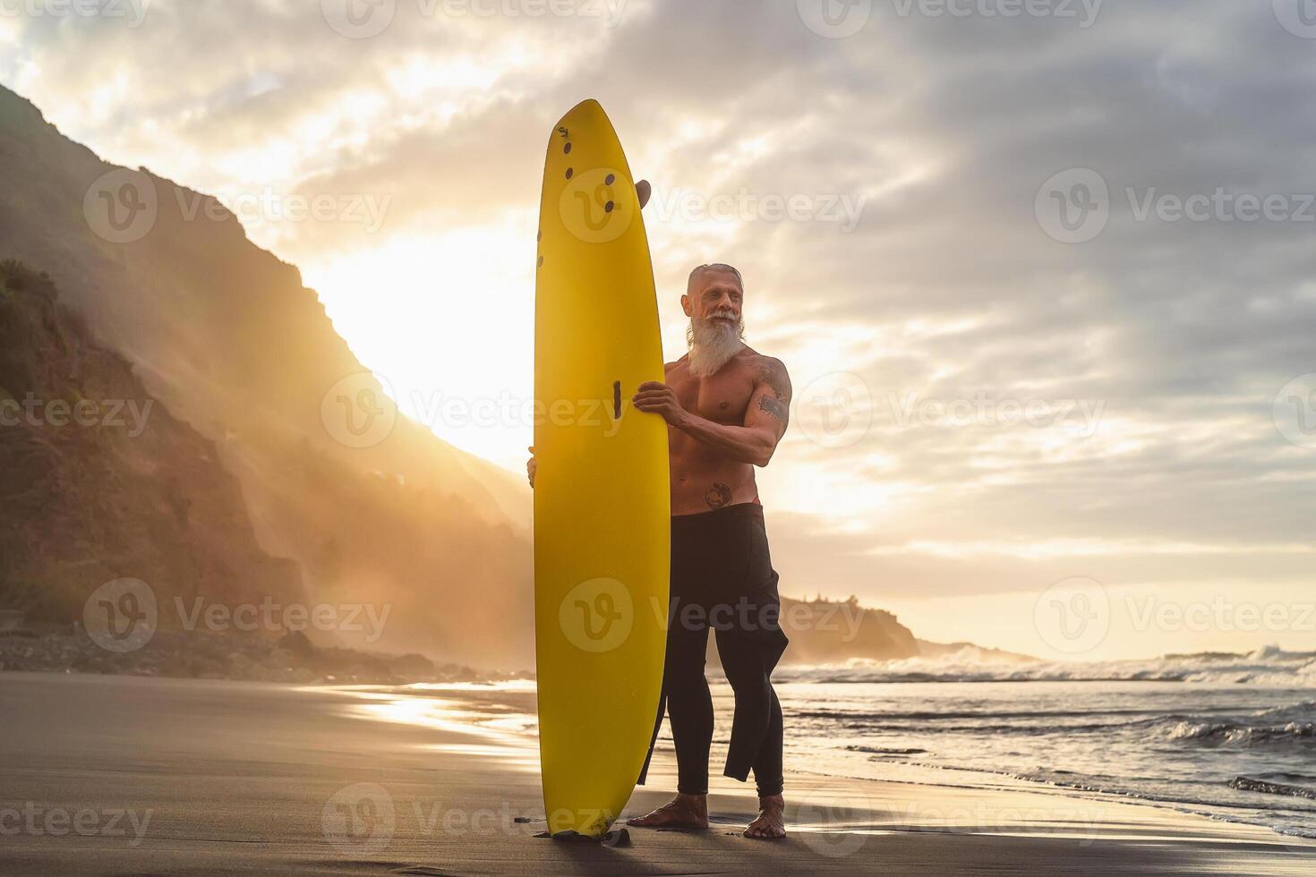 Happy fit senior having fun surfing at sunset time - Sporty bearded man training with surfboard on the beach - Active elderly people lifestyle and extreme sport concept photo