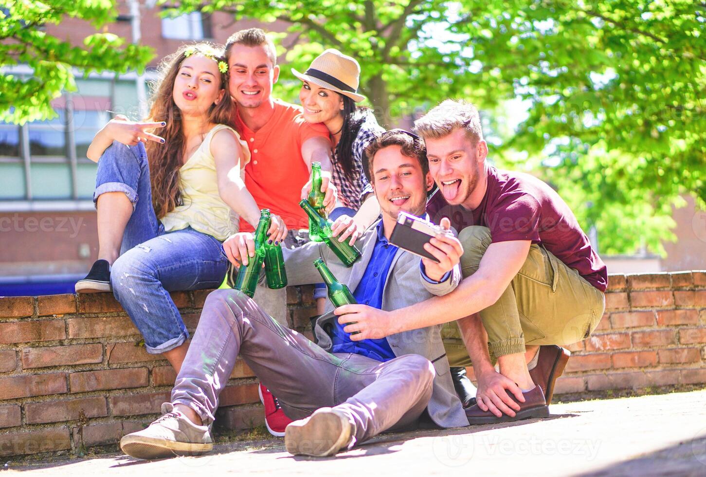 Group of happy friends drinking beers and taking selfie with a vintage camera outdoor - Young people making photos while toasting and cheering bottles of beer in the city - Friendship, youth concept