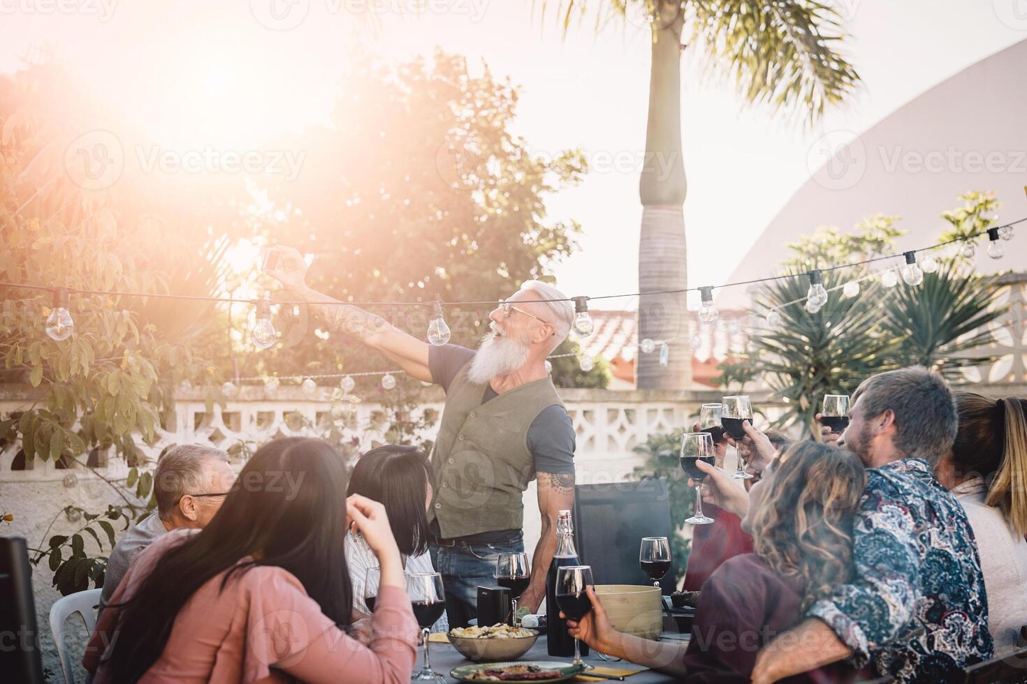 Happy family taking selfie with mobile smartphone camera at dinner party outdoor - People with different ages and ethnicity eating together on patio - food and drink, retired and young people concept photo