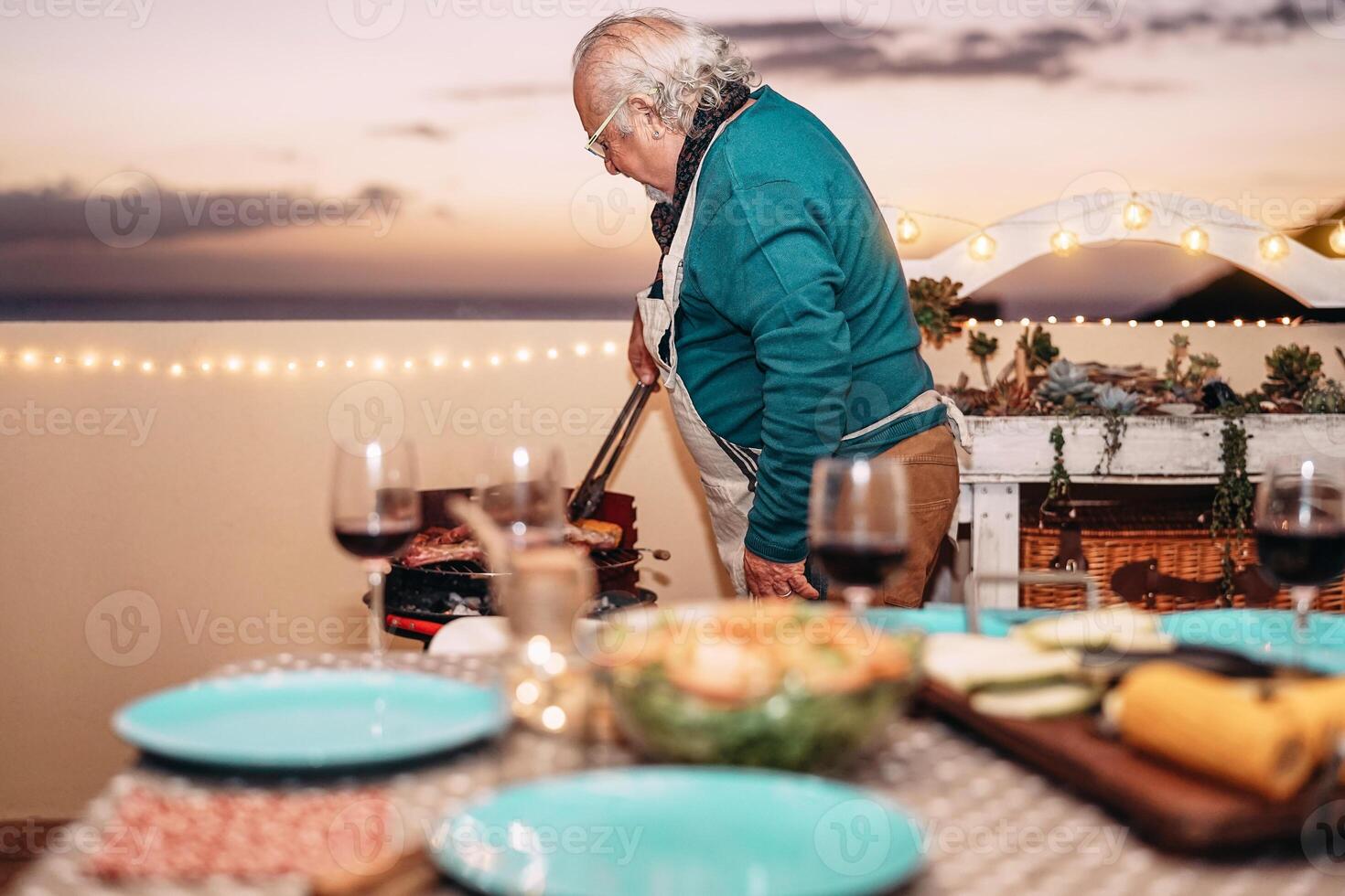 Senior man grilling meat at barbecue dinner on terrace - Grandfather cookin for his family on rooftop - Concept of dining bbq and eating together outdoor photo