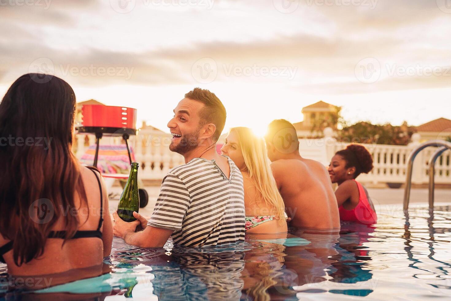 Happy friends making a pool party drinking champagne in the poolside - Young people in swimsuits having fun at barbecue dinner in luxury tropical resort at sunset - Youth holidays lifestyle concept photo