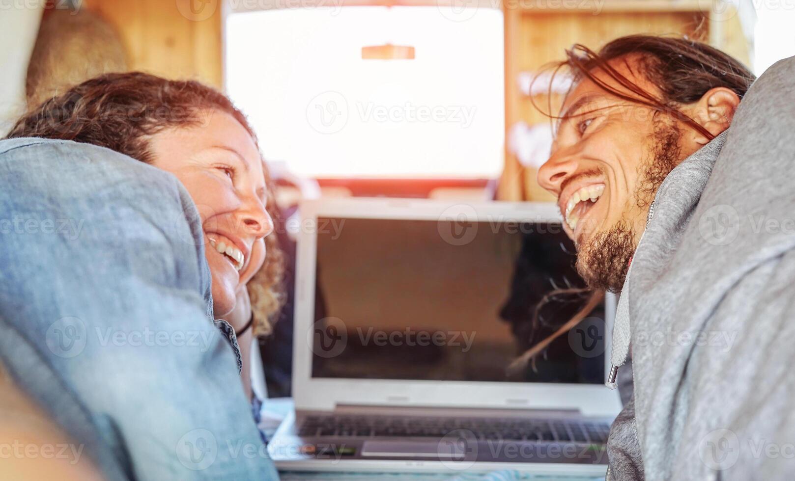 Happy man and woman lookin each other and using their computer in caravan - Travel couple tender moment during their journey on vintage minivan with wood interior - Vacation, love, technology concept photo
