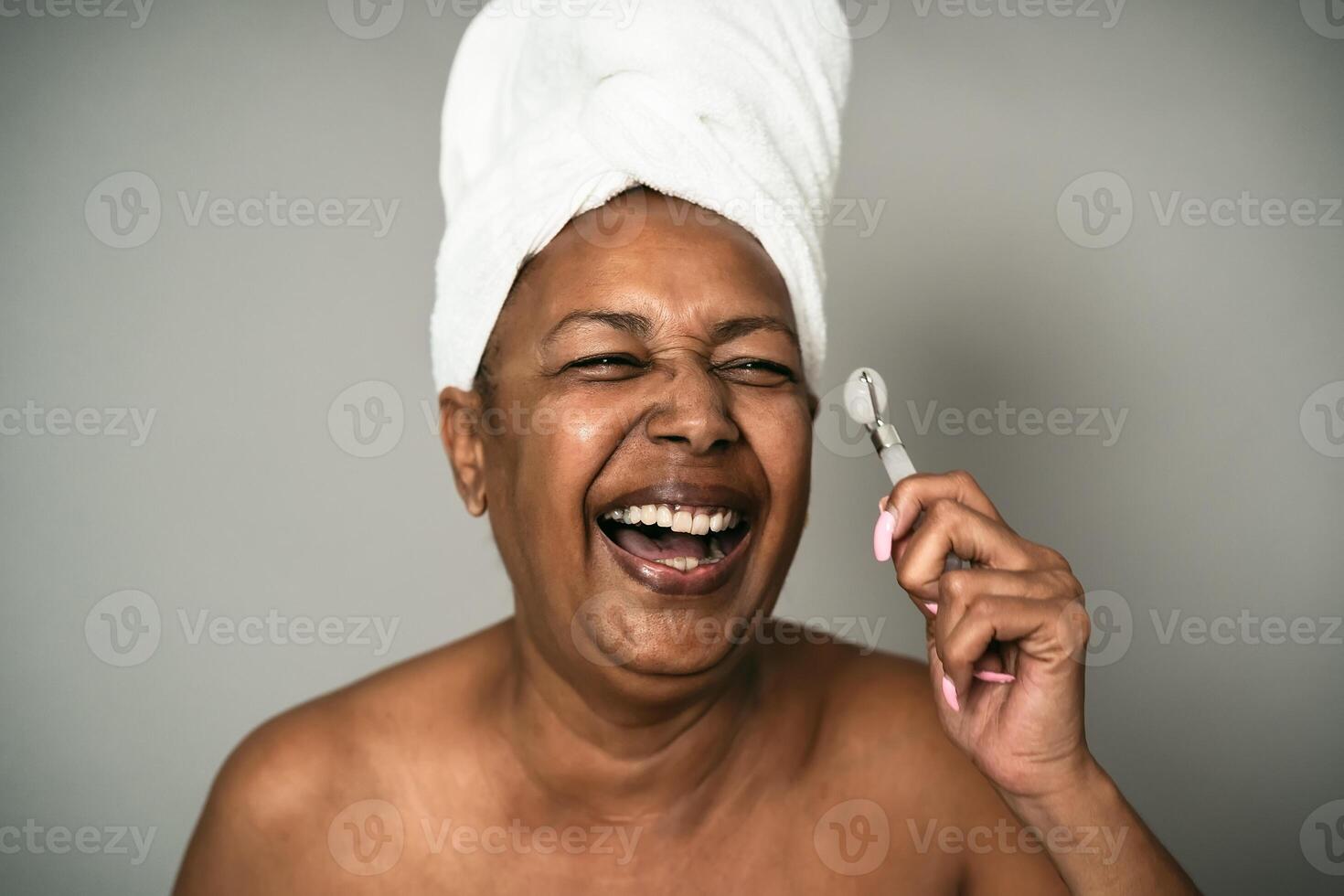 Happy Afro senior woman using jade roller during skin care spa day - People wellness lifestyle concept photo