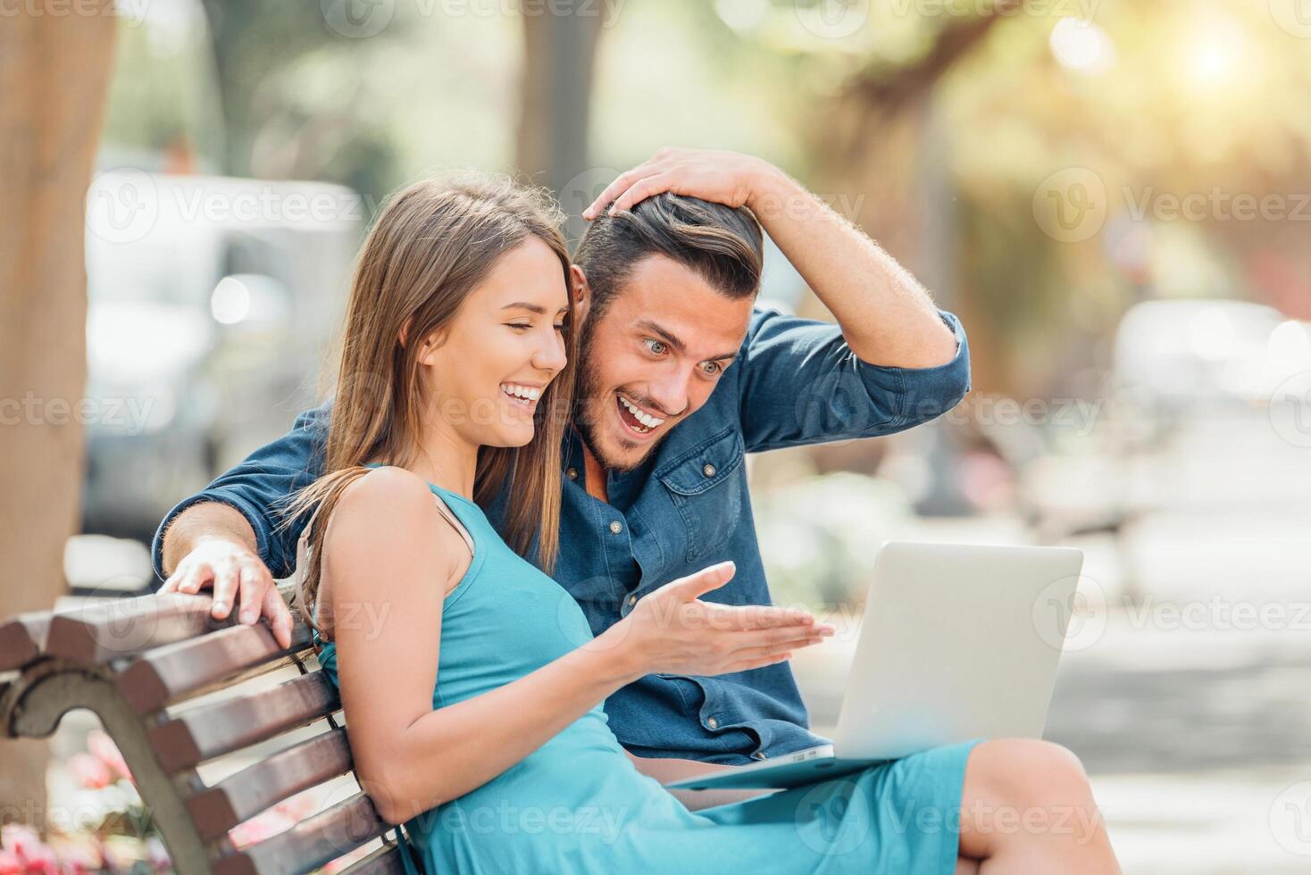 Happy young couple using laptop computer sitting on bench in city outdoor - Handsome man surprised to what his girlfriend is showing to him - Concept of relationship and people addicted to technology photo