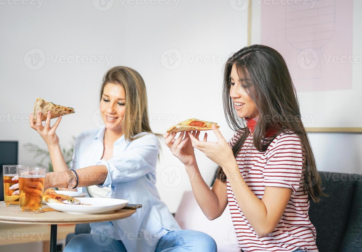 joven mujer comiendo rápido comida Pizza y Bebiendo cerveza a hogar - contento amigos disfrutando un cena sentado en sofá en el vivo habitación en su Departamento - personas comida concepto foto