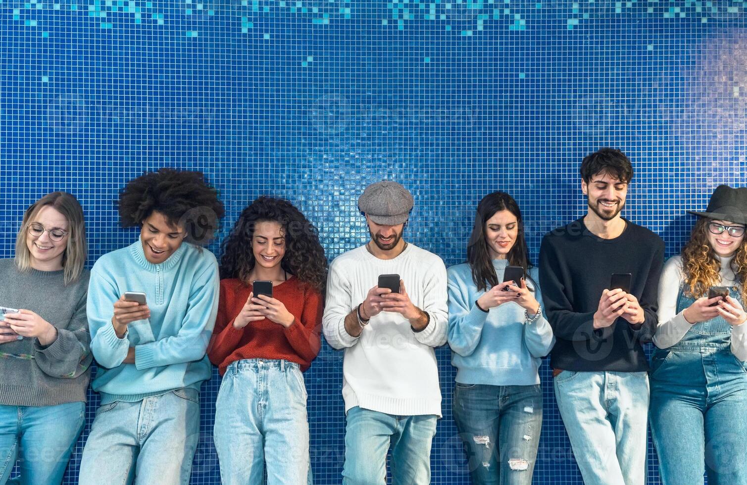 Young people using mobile smartphone while standing against blue background - Youth millennial addicted to new technology social media app concept photo