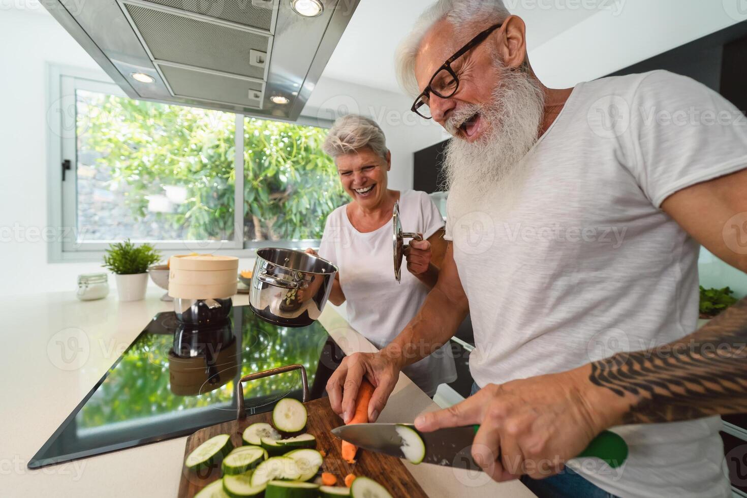 Happy senior couple having fun cooking together at home - Elderly people preparing health lunch in modern kitchen - Retired lifestyle family time and food nutrition concept photo