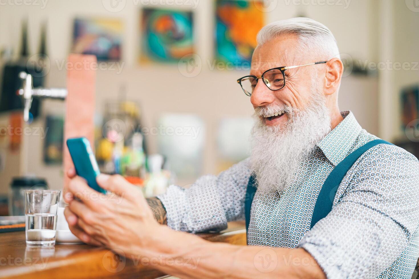 Hipster man using mobile phone while sitting in a bar - Senior male watching on new trends smartphone apps for social media - Technology and people lifestyle concept photo