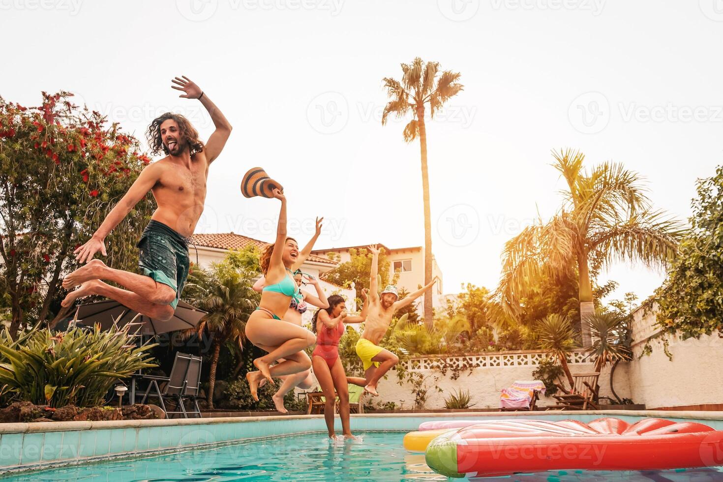 Group happy friends jumping in pool at sunset time - Crazy young people having fun making party in exclusive tropical house - Summer holidays vacation and youth culture lifestyle concept photo