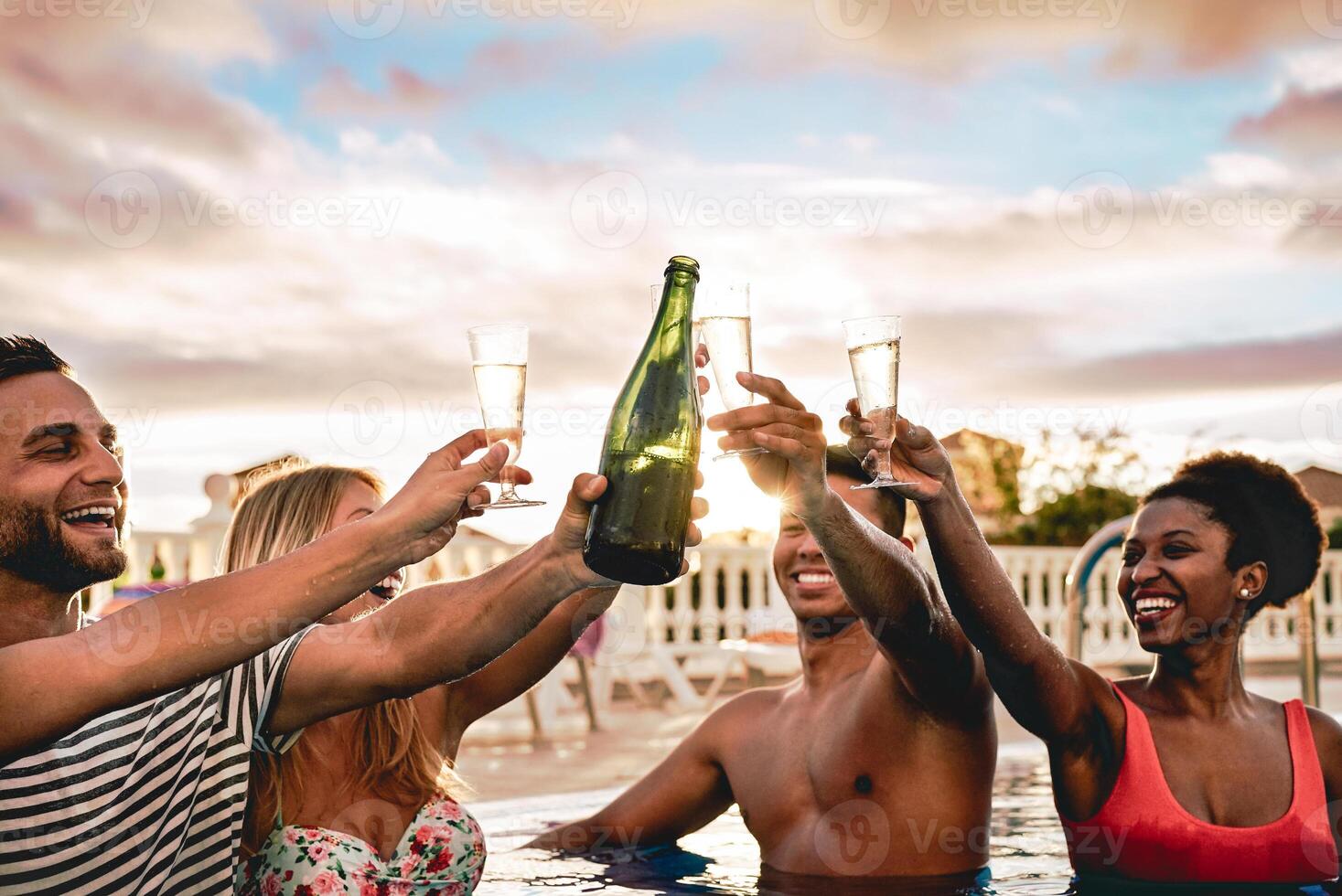 Happy friends doing pool party toasting champagne at sunset - Young people having fun drinking sparkling wine in luxury tropical beach resort - Holidays, vacation, summer and youth lifestyle concept photo