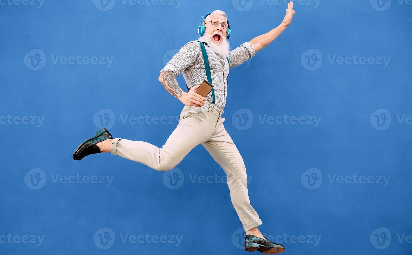 Senior crazy man jumping while listening music with wireless headphones - Hipster old guy having fun dancing outdoor - People happiness and elderly technology addicted concept - Blue background photo