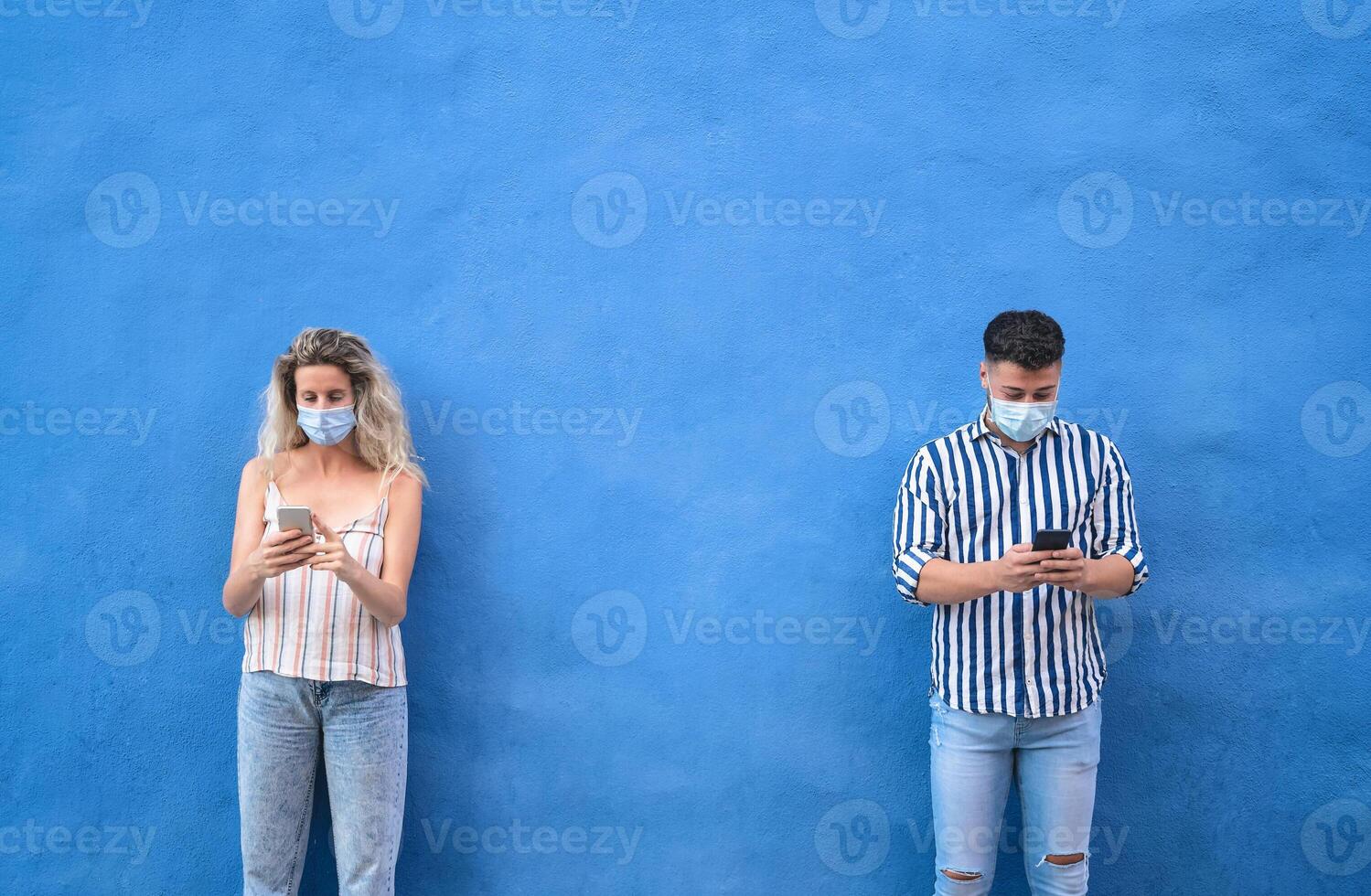 Young people wearing surgical mask using mobile phone keeping social distance - Millennial friends watching media trends on smartphones during corona virus outbreak - Youth and technology concept photo