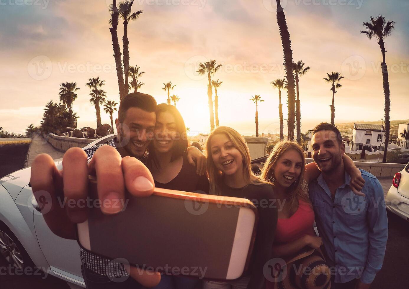 Happy friends standing in front of convertible car and taking selfie with mobile phone camera - Young millennial people having fun on roadtrip in tropical destination - Travel and technology concept photo