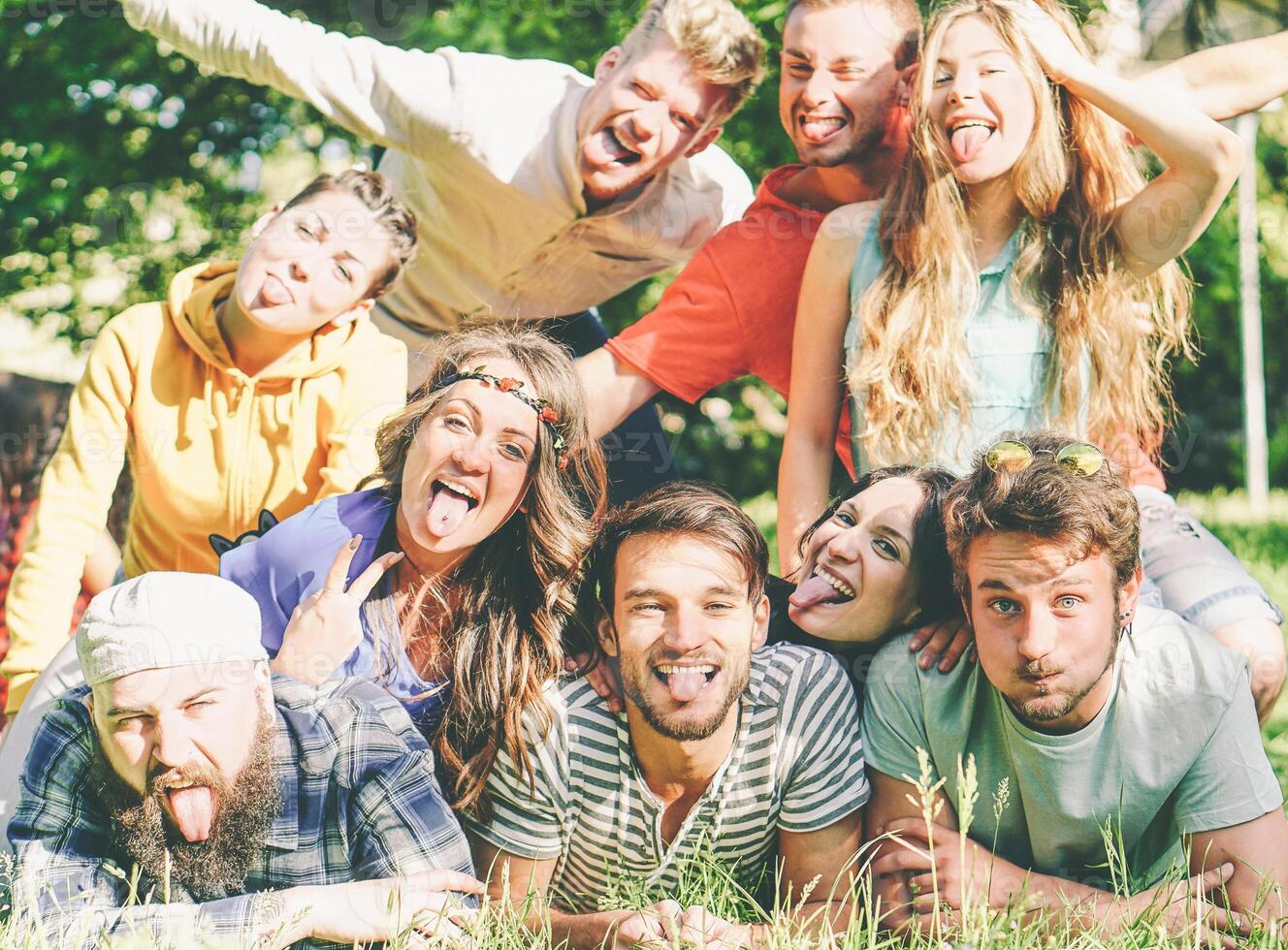 Group of friends having fun together lying on grass while making a self portrait with silly faces - Young happy people taking photo of group at picnic camping park - Friendship and lifestyle concept