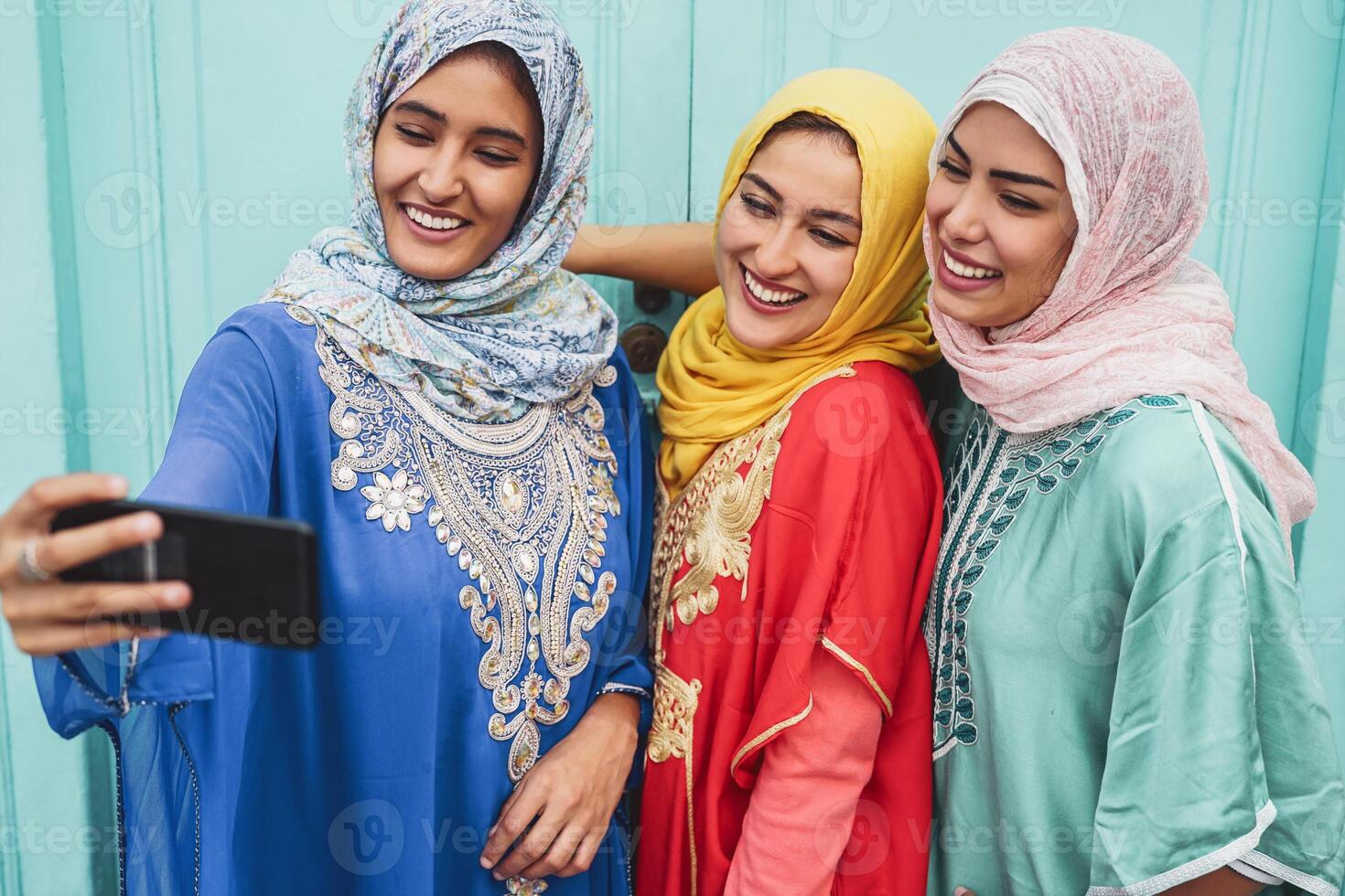 Happy Muslim girls taking selfie with mobile smartphone camera outdoor - Arabian young friends making photo for social media network - Religion culture and technology concept