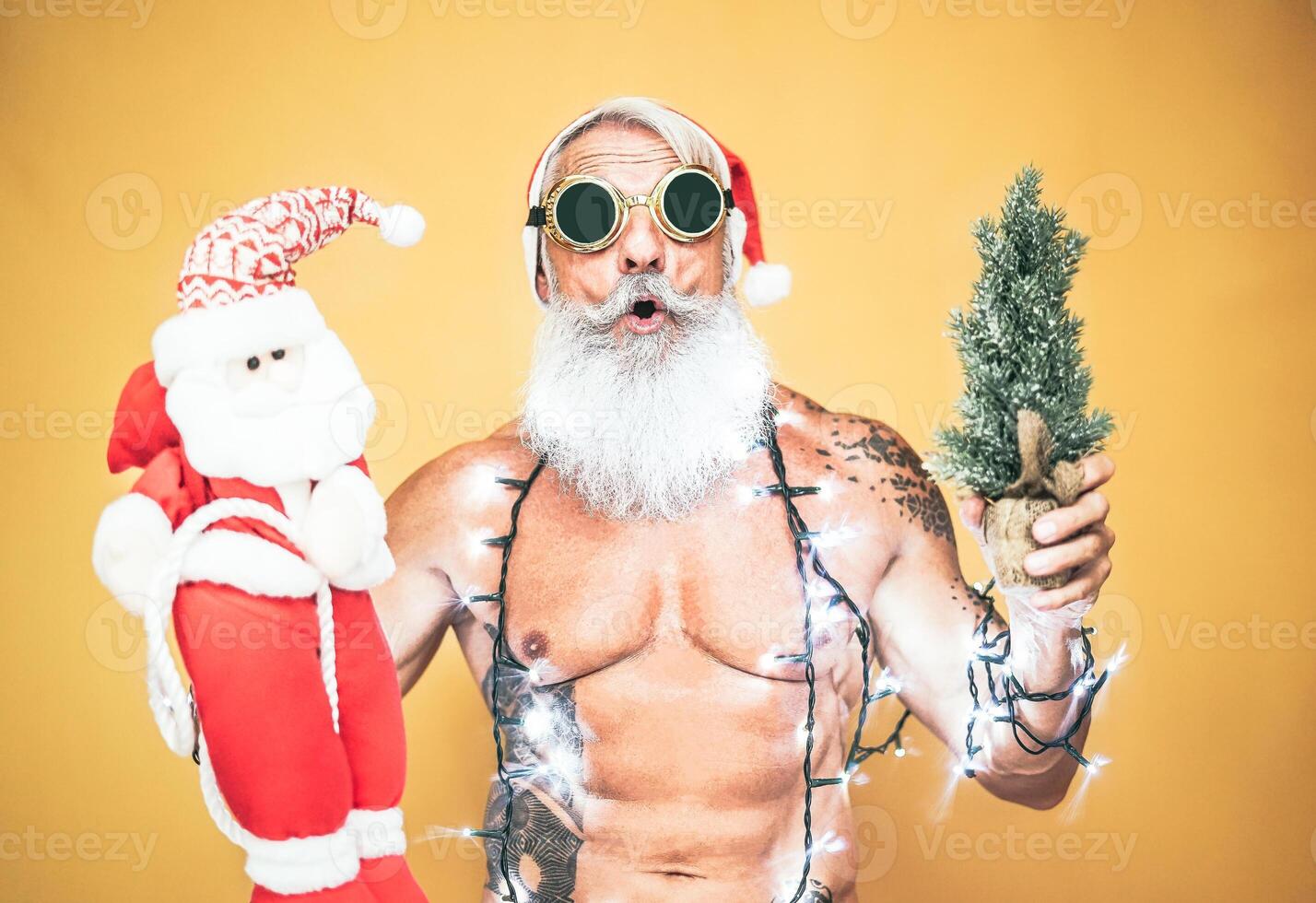 Happy fit santa claus equiped with white christmas lights - Trendy beard hipster senior holding a mini santa claus puppet and xmas tree - Celebration and holidays concept photo