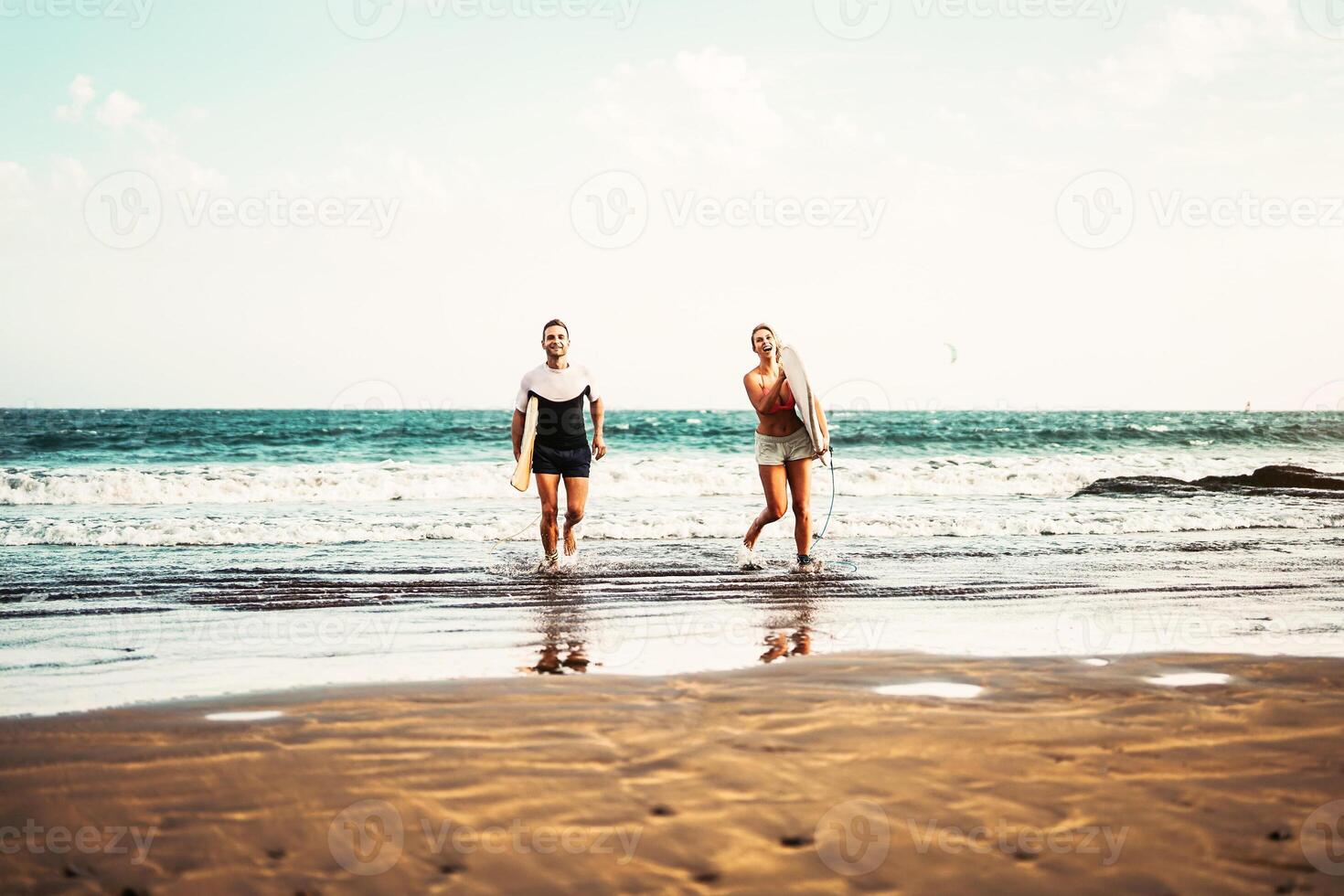 Happy surfers couple running out water after ride on waves - Young people having fun surfing together - Extreme sport and youth culture lifestyle concept photo