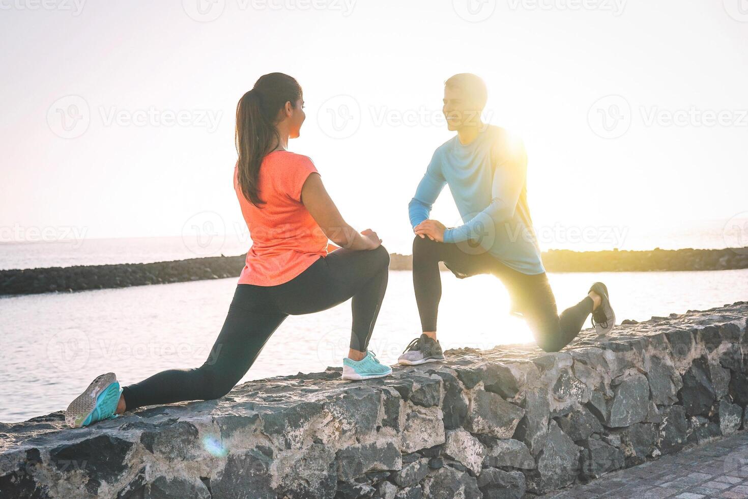 joven salud Pareja extensión piernas siguiente a el playa a puesta de sol - contento juguetón amantes rutina de ejercicio juntos en pie en frente cada otro y sonriente - relación, estilo de vida, deporte concepto foto