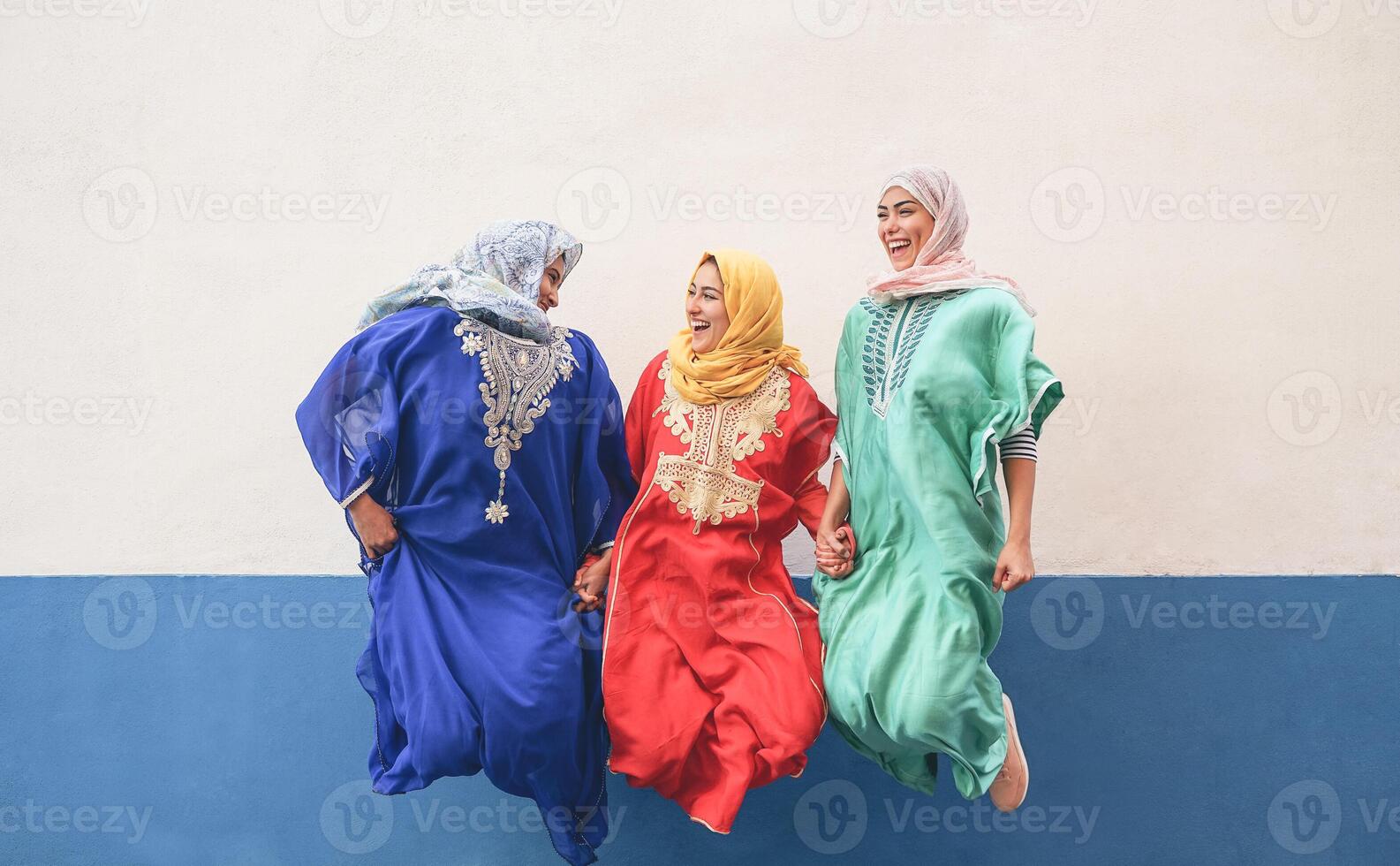 Happy Muslim women having fun jumping and laughing together outdoor - People lifestyle and culture religion concept - White and blue background photo