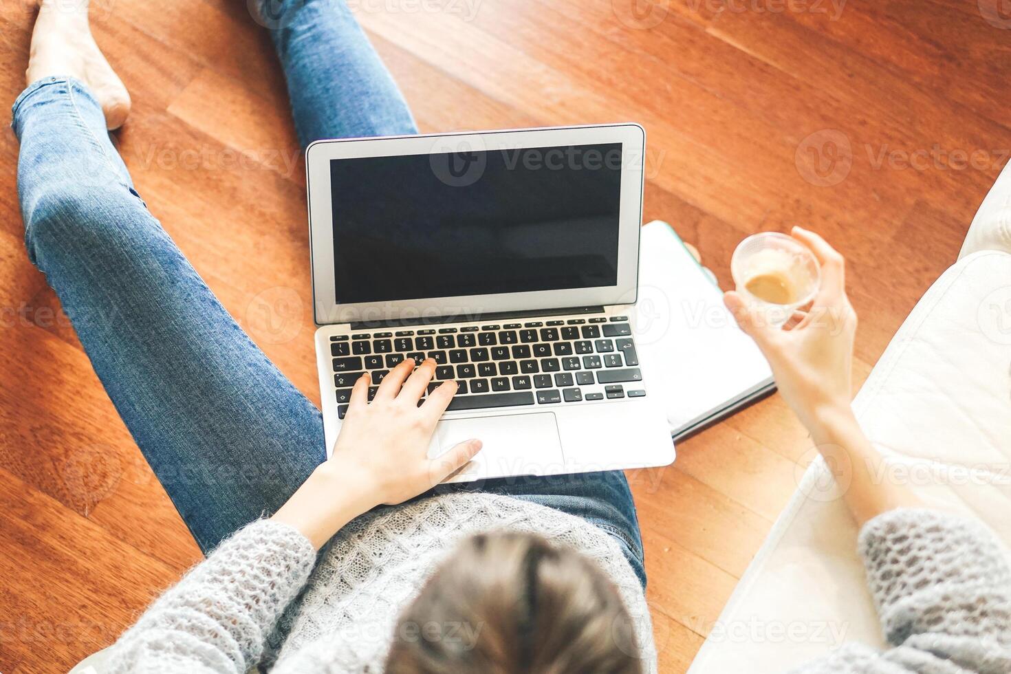 Top view of a young student using her computer while sitting on the floor under sofa holding coffee in her hand - Woman studying on her laptop while drinking coffee in living room - Focus on notebook photo
