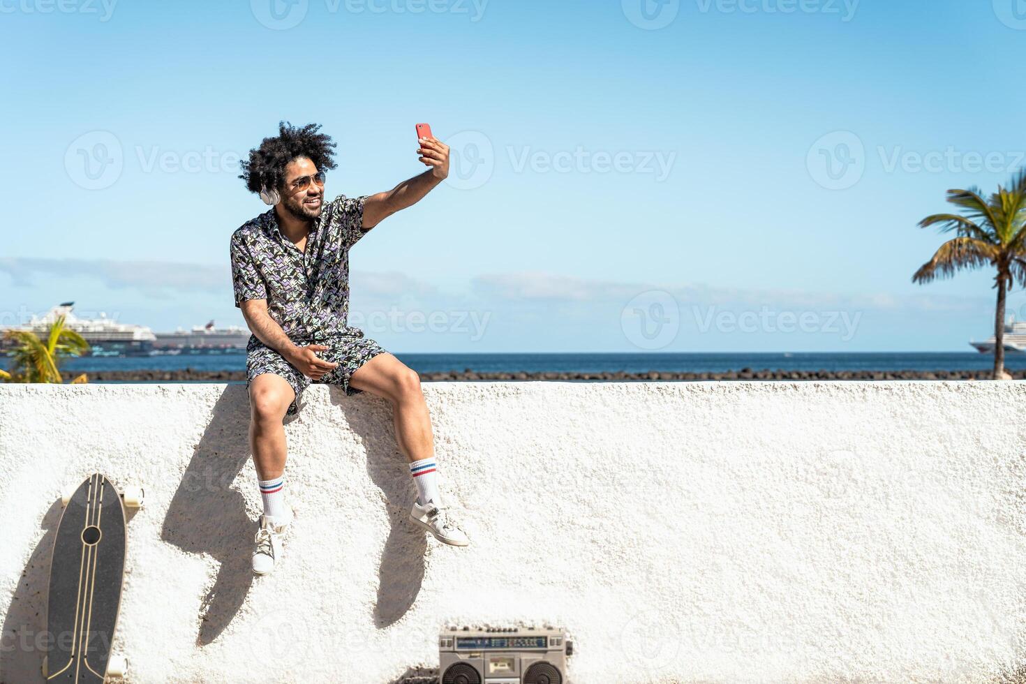 Young Latin man having fun taking selfie with mobile smartphone while listening music with headphones and boombox during summer vacations photo