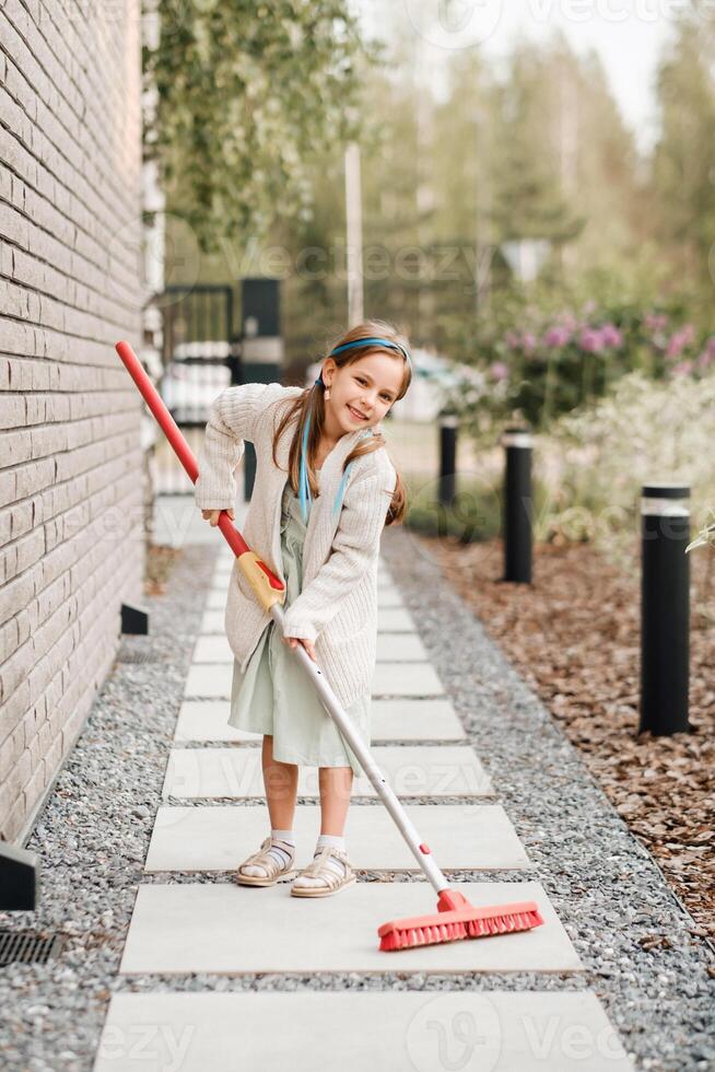 un pequeño niña con un cepillo limpia un camino en el calle en el patio foto