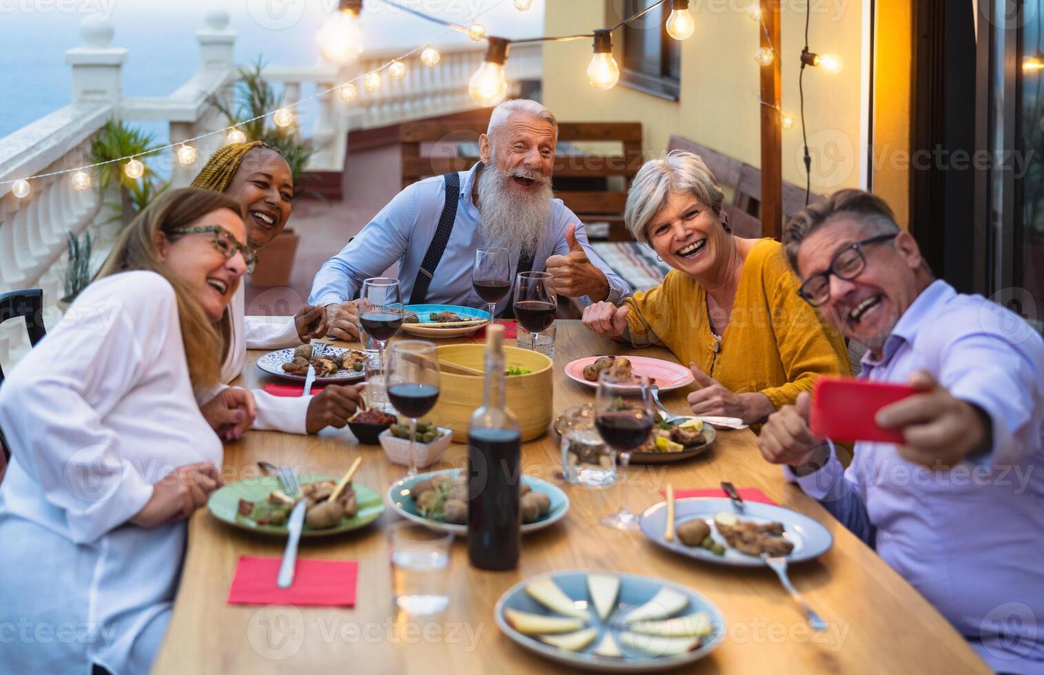 Happy multiracial senior friends having fun dining together while taking selfie with mobile smartphone on house patio - Elderly lifestyle people and food concept photo