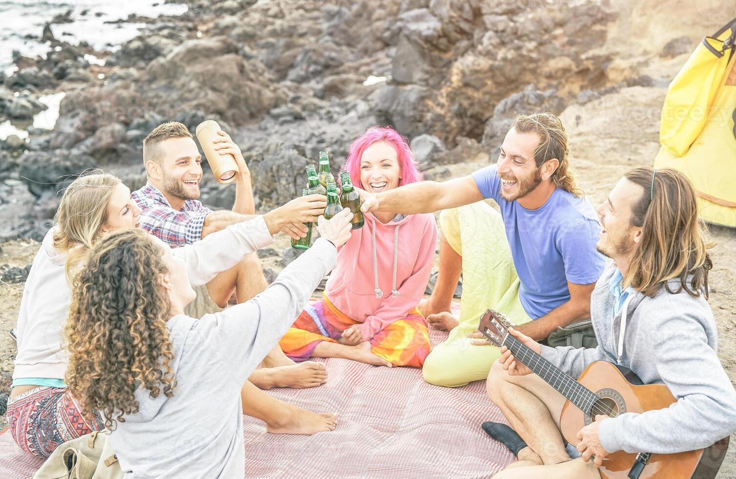 Group of happy friends toasting beers and playing guitar camping with tent on the beach - Travel people having fun listening music and drinking alcohol outdoor - Vacation, youth lifestyle concept photo