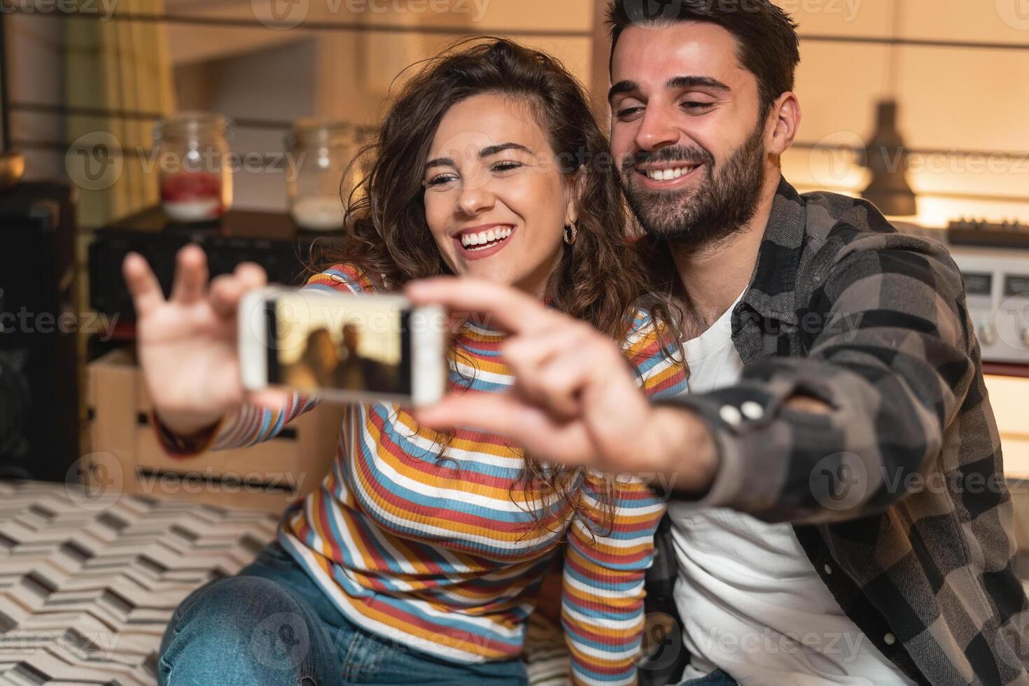 Happy couple taking selfie with mobile smartphone in living room at home - Young lovers having fun with new trendy social media apps - Love relationship technology concept photo