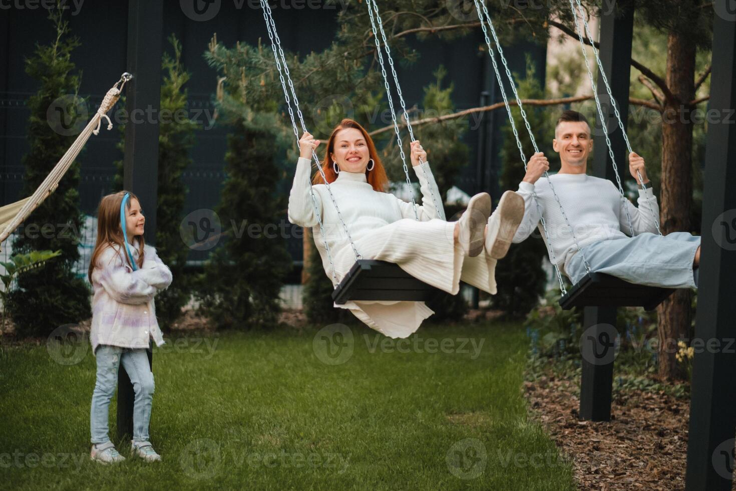 Mom and dad are riding on a swing and there is a daughter standing next to them. The family is resting on a swing photo