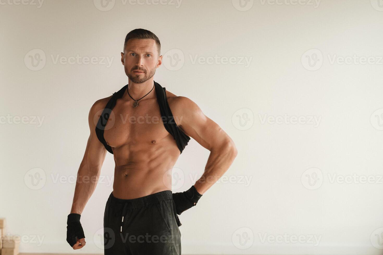 An adult muscular man with a naked torso stands in a room on a light background photo