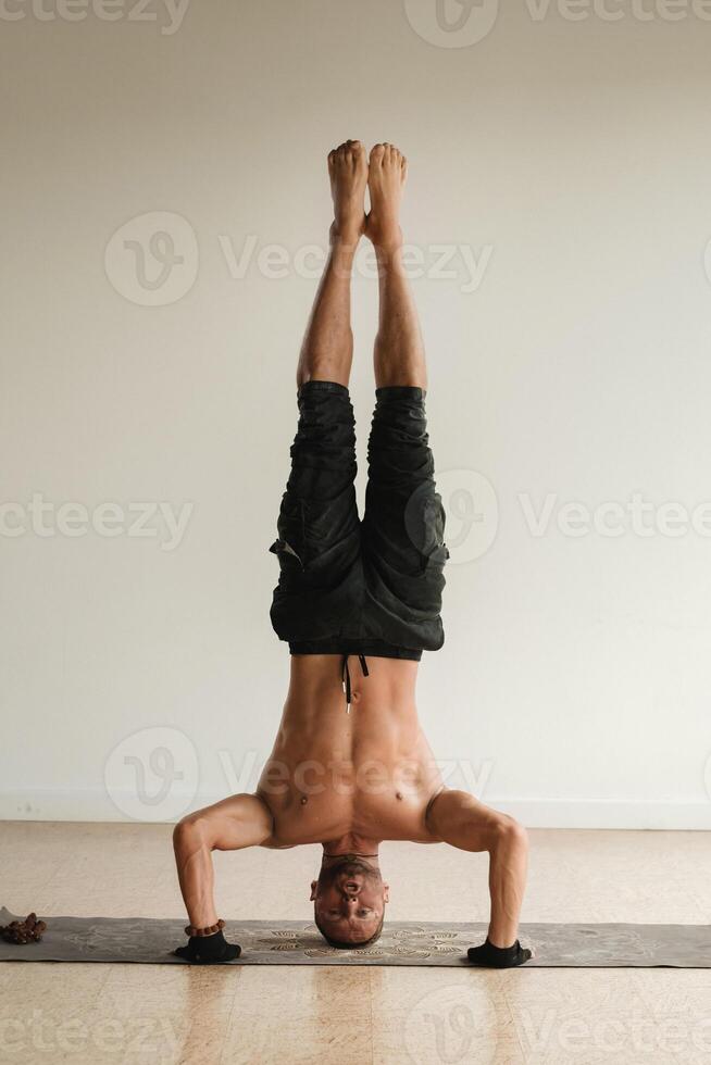 a man with a naked torso does yoga standing on his head indoors. Fitness Trainer photo