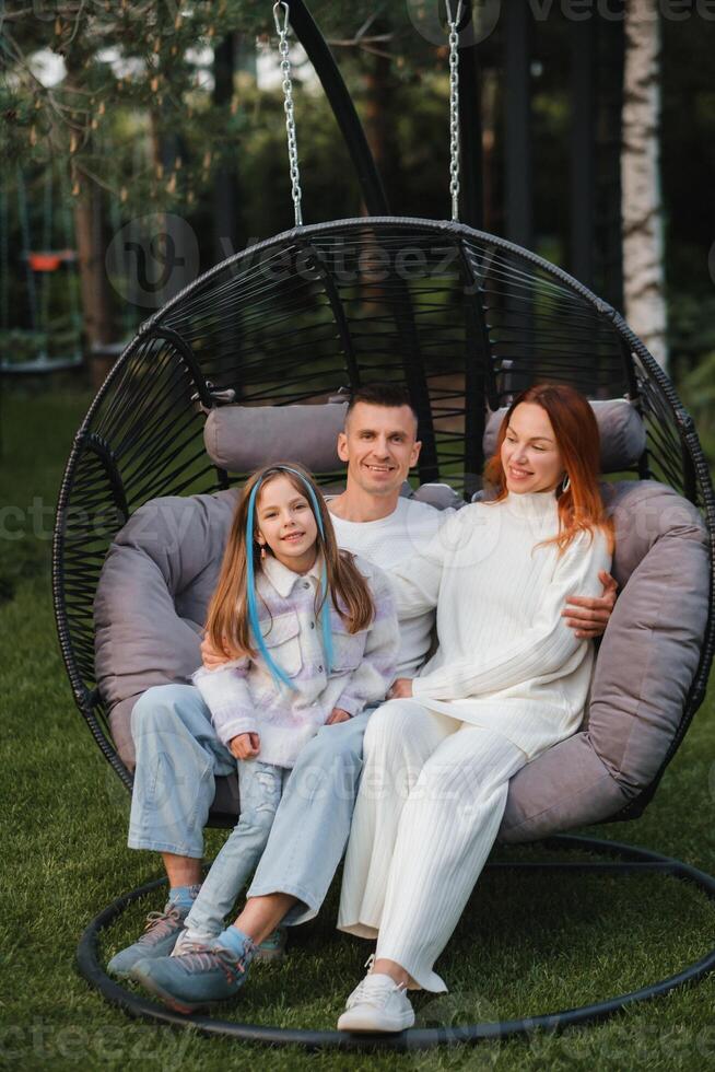 un contento familia es sentado en un hamaca en el césped cerca el casa foto