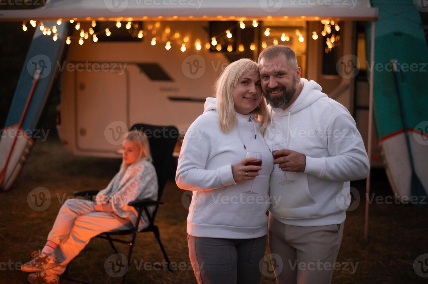 A married couple with glasses of wine stands against the background of a motorhome and rests together by the campfire. Evening family vacation photo