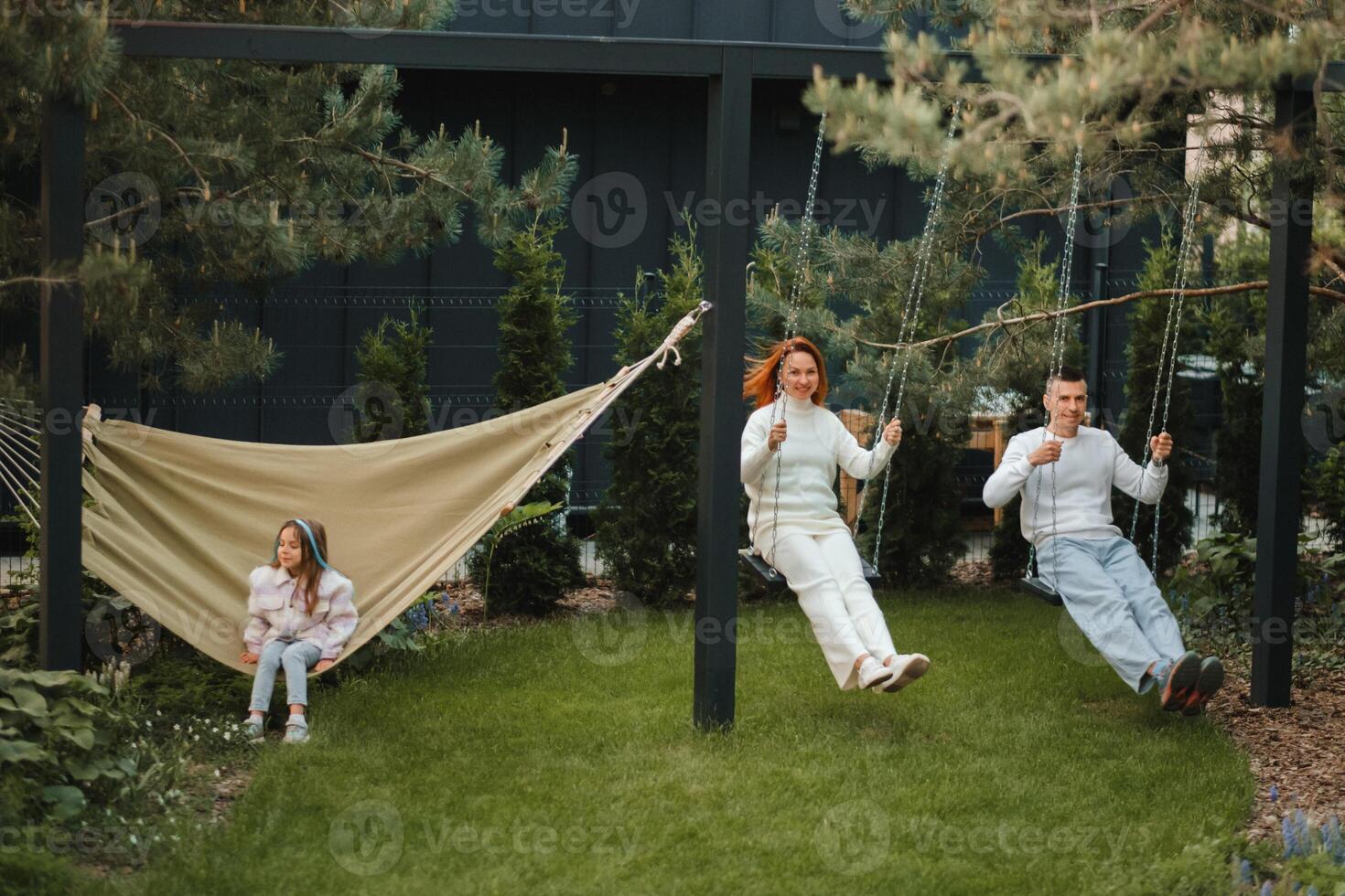 Mom and dad are riding on a swing, and their daughter is on a hammock next to them. The family is resting on a swing photo