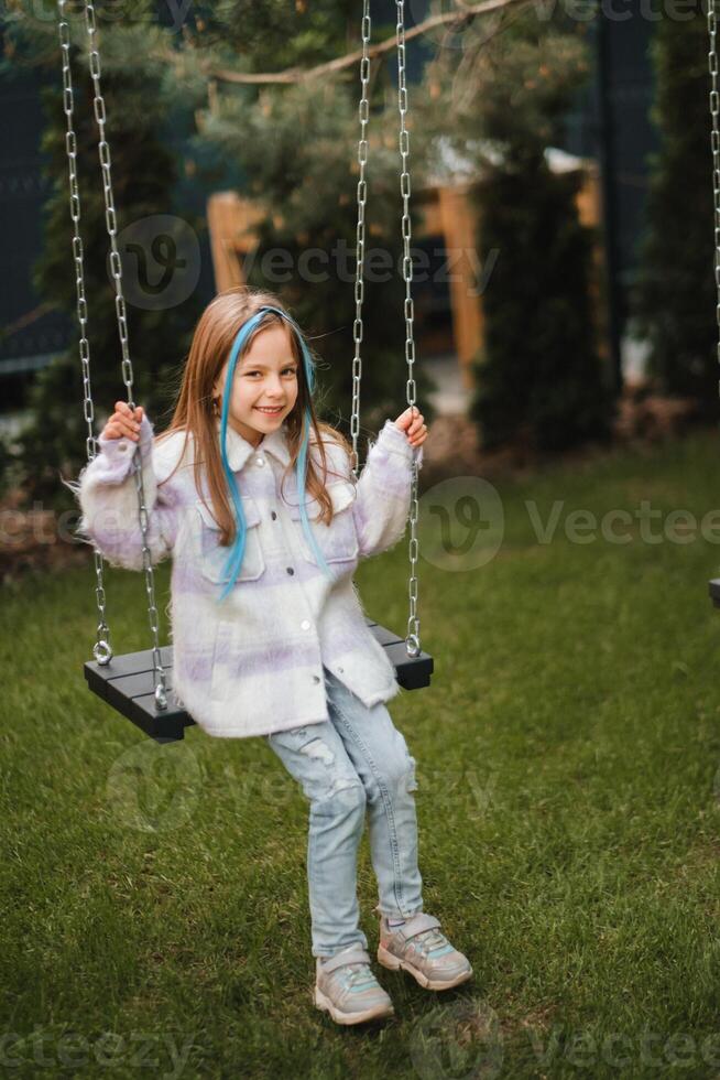 A beautiful girl with long hair in a jacket rides on a swing in the evening on the street photo