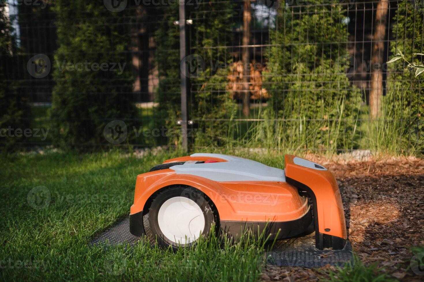 An orange robotic lawn mower stands on the base and is charged from electricity in the courtyard photo