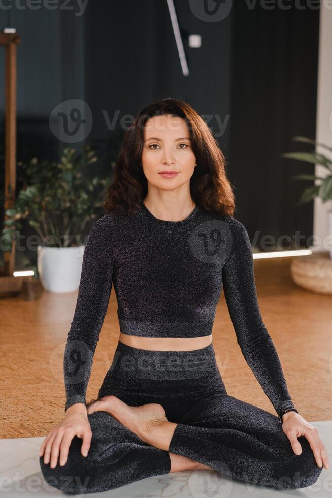 A woman in a black tracksuit sits on a yoga mat in a lotus position in the gym photo