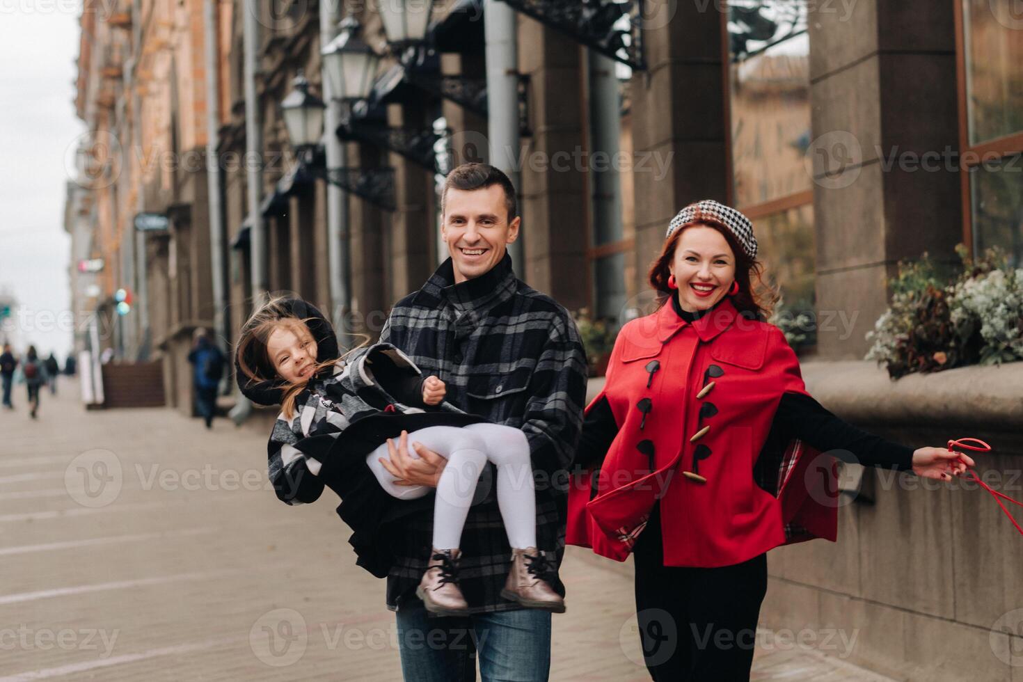 A stylish family of three people are walking in the city in the fall and dad is holding his daughter in his arms photo