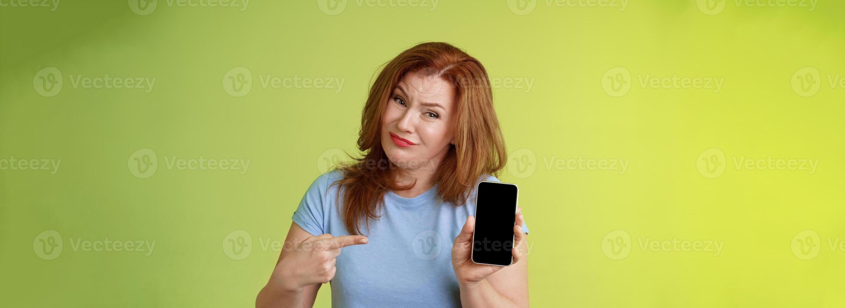 Seriously it awful. Displeased disappointed redhead mature female tilt head cringe grimacing reluctant pointing smartphone blank display index finger showing bad photograph share negative opinion photo