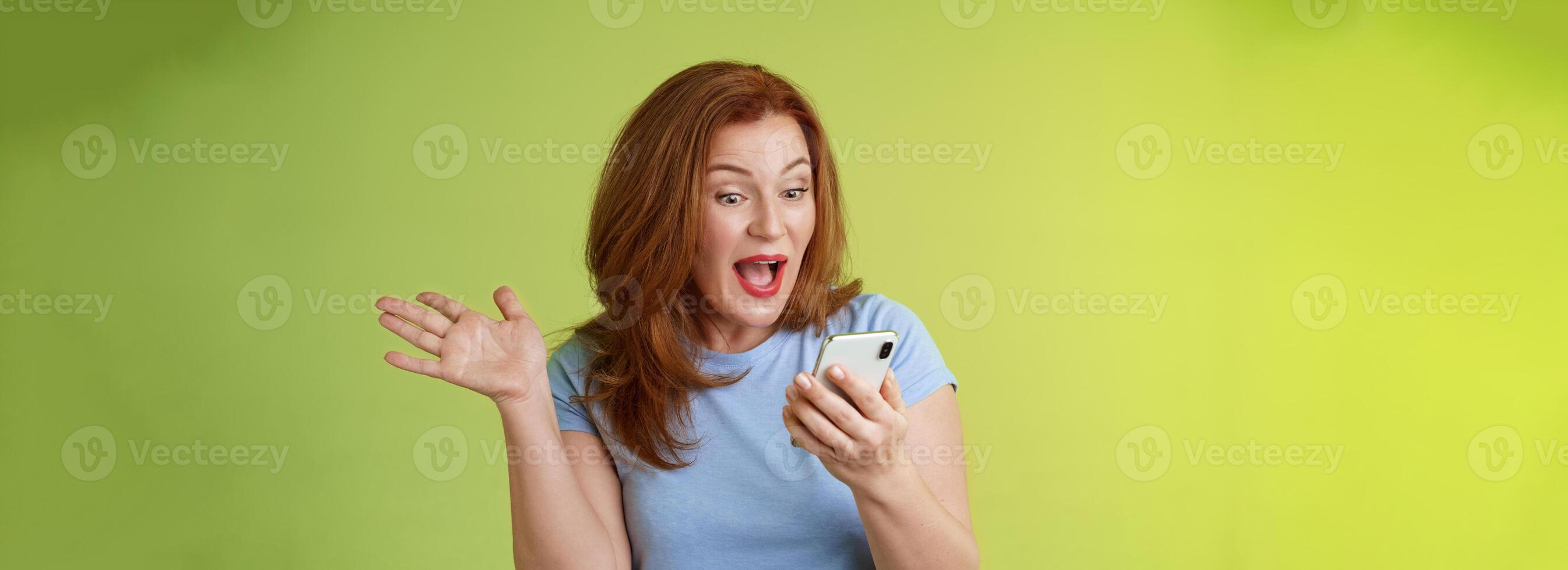 Surprised glad middle-aged pretty redhead woman look smartphone raise hand celebratory excitement open mouth reacting impressed gladly read good excellent news look mobile screen green background photo