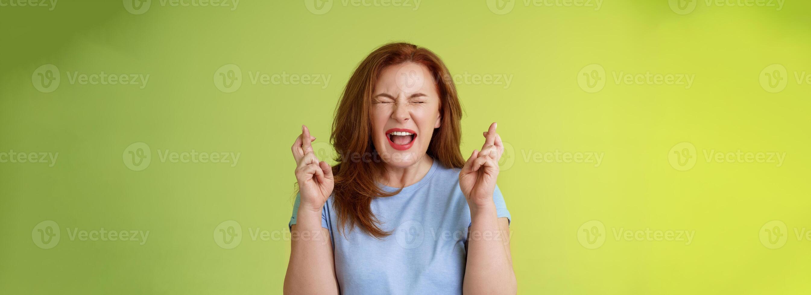 Woman wants win badly. Enthusiastic lucky redhead middle-aged 50s female pleading implore god make dream come true cross fingers good luck wishing closed eyes open mouth excitement green background photo