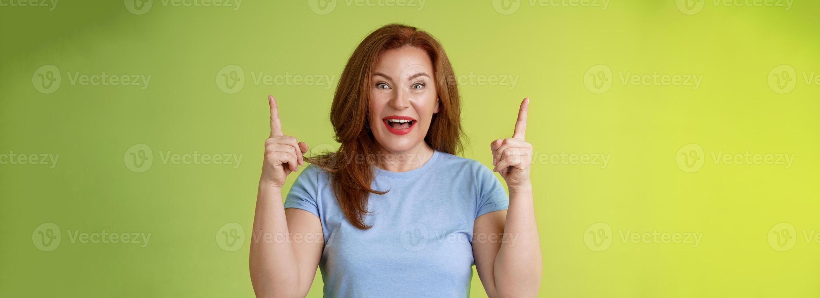 Impressed enthusiastic happy cheerful middle-aged mature woman red hair smiling astonished look admiration joy pointing up raised index fingers introduce impressive stunning offer green background photo