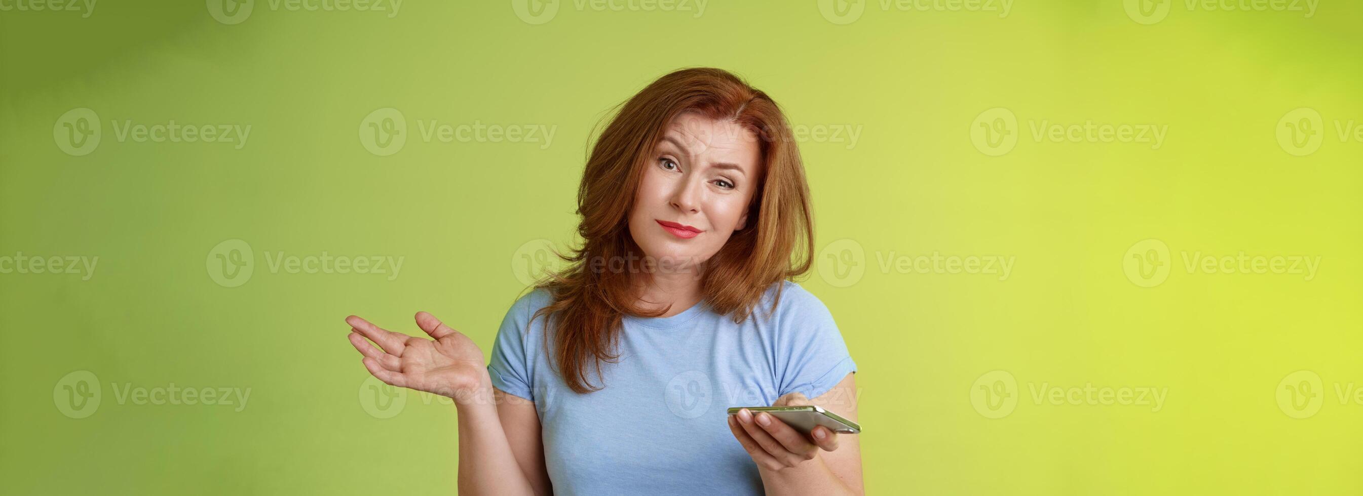 Well meh. Indifferent careless hesitant redhead middle-aged woman mature red female shrugging hold smartphone smirk bored uninterested hold hand aside apathetic attitude green background photo